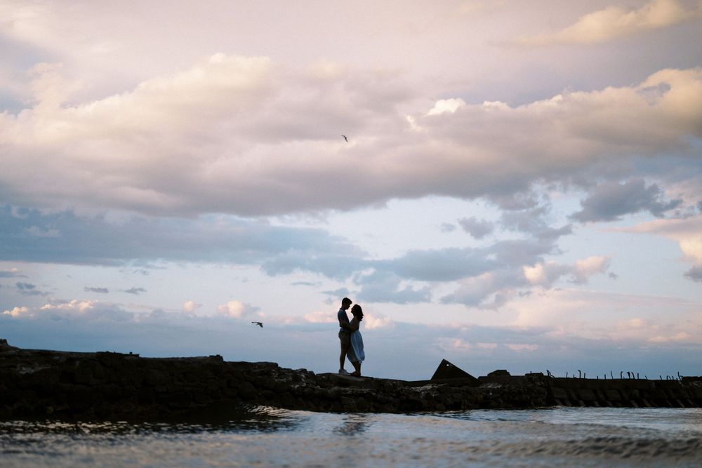 A silhouetted figure stands on a rocky beach at sunset with dramatic clouds in the background.