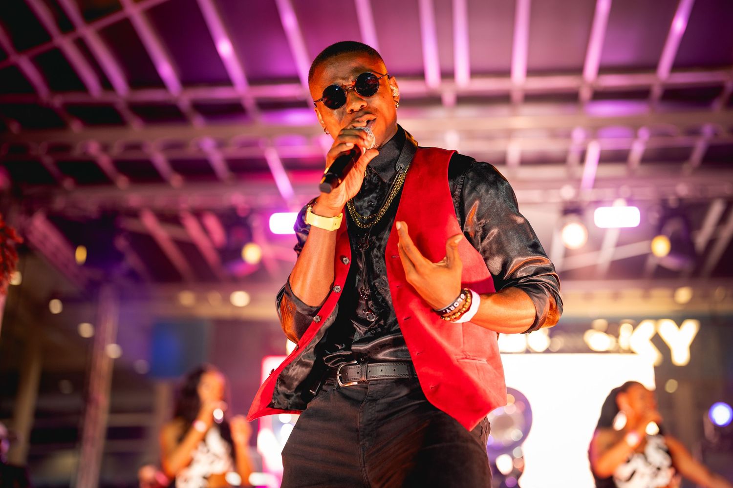 Performer wearing sunglasses and a red vest sings passionately on stage with dramatic purple lighting in the background.
