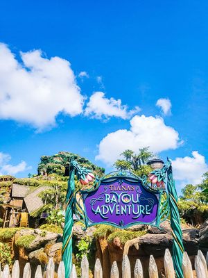Entrance sign for the Bayou Adventure ride featuring tropical foliage and blue skies at a theme park.