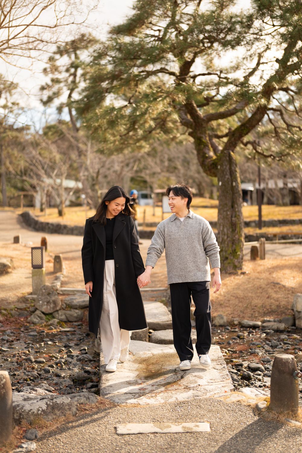 Smiling couple holding hands walking on stone path in sunlit Japanese park with large pine tree.