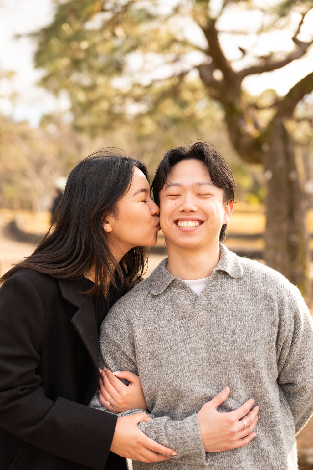 Woman kissing smiling mans cheek while embracing him outdoors in sunny Japanese park with trees.