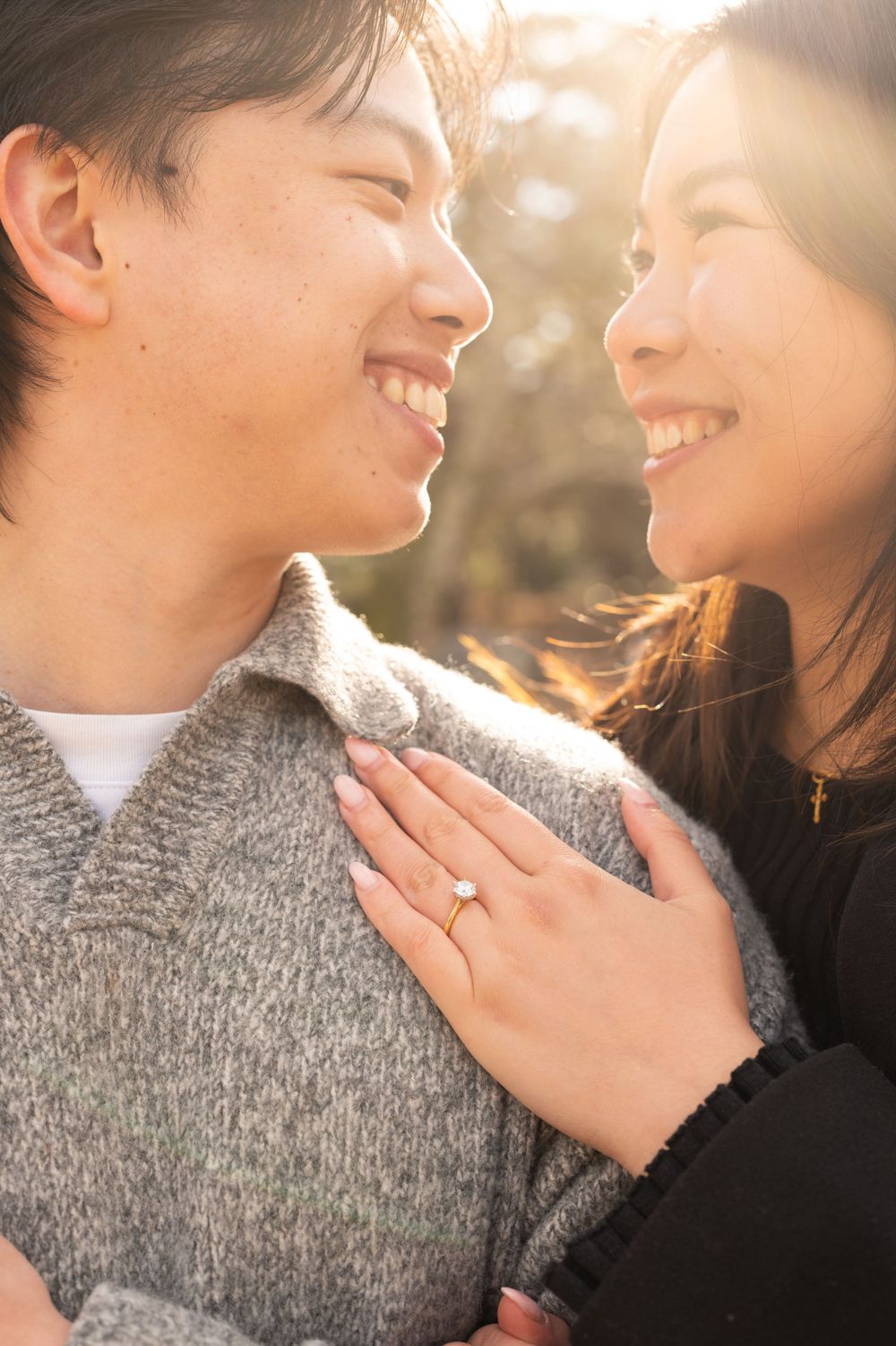 Couple smiling at each other with engagement ring visible, backlit by golden sunlight in an outdoor setting.