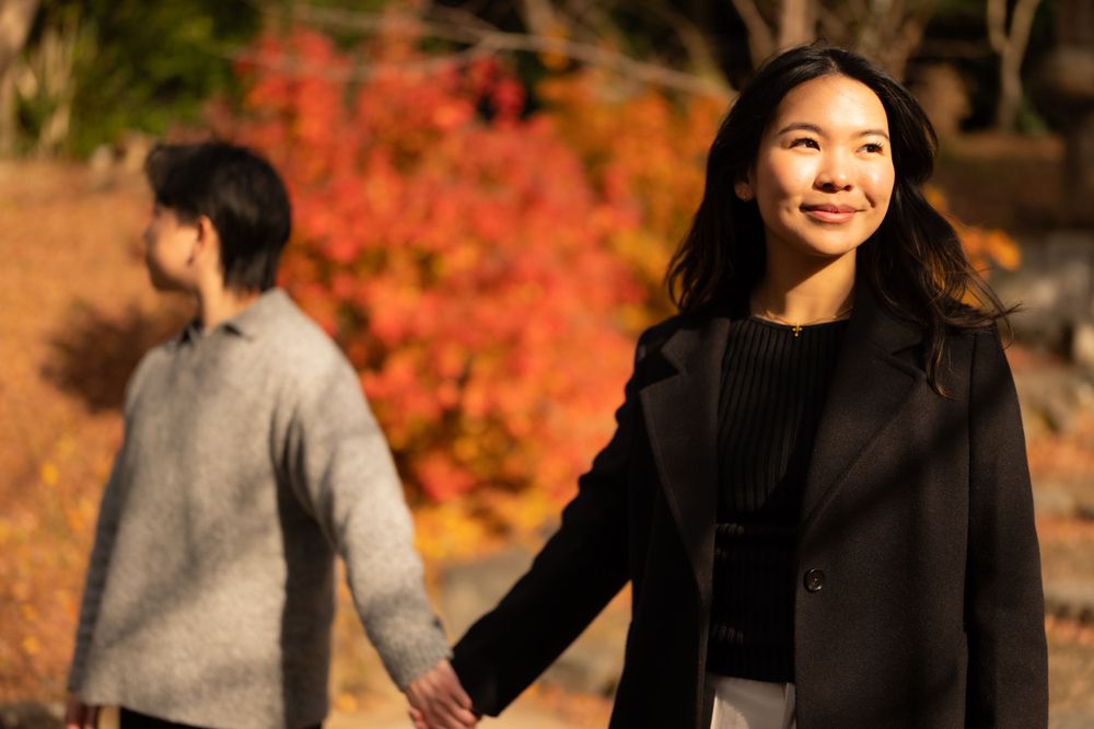 Woman in black coat smiling while holding hands with partner amid vibrant autumn foliage outdoors.