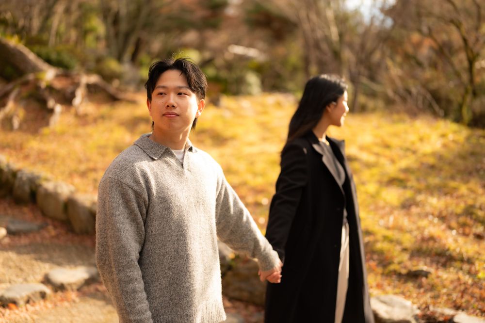 Man in gray sweater glancing back while holding hands with woman on a scenic autumn park path.