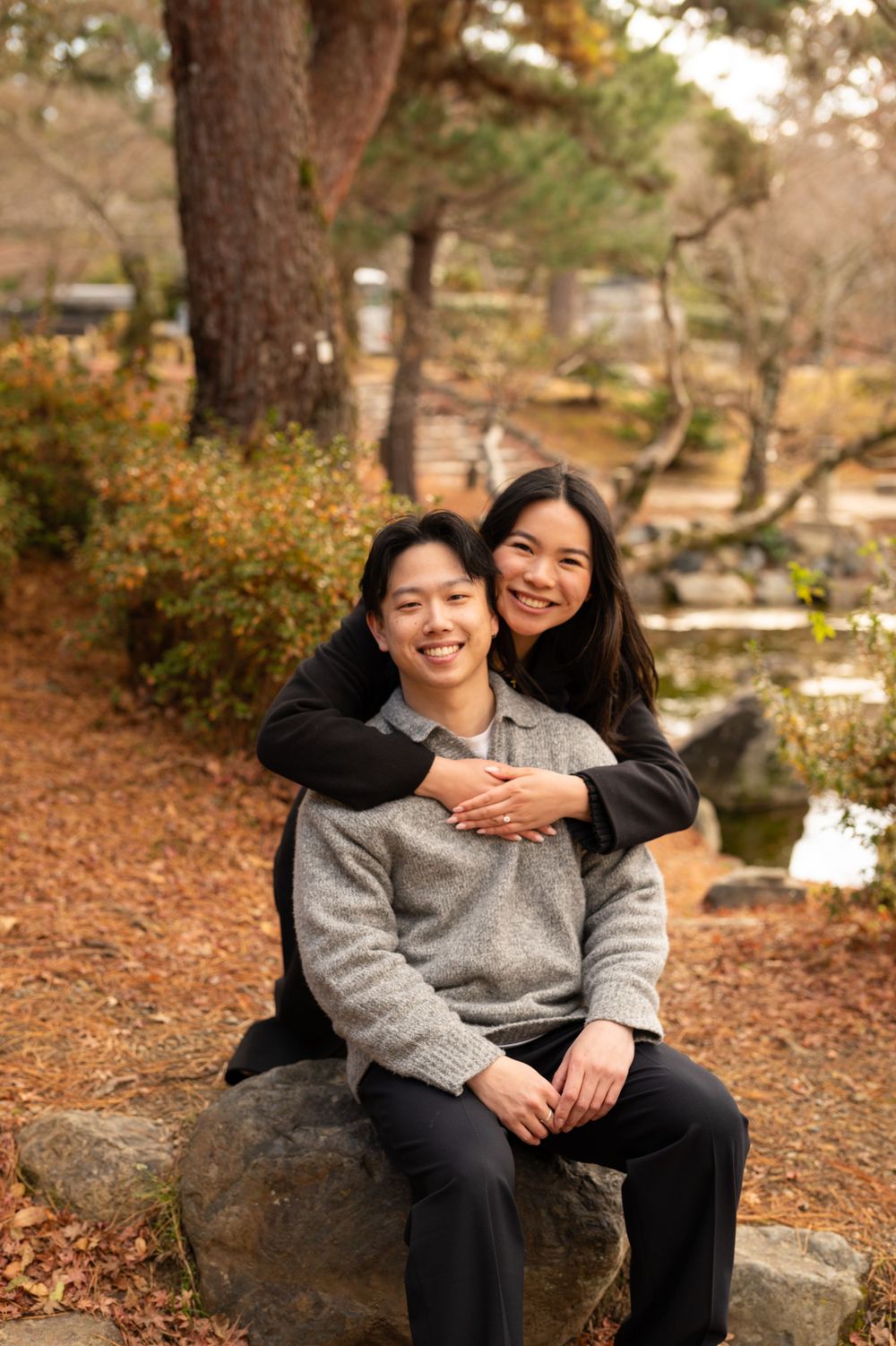 Smiling couple posing in a Japanese garden, woman embracing man from behind on a stone seat.