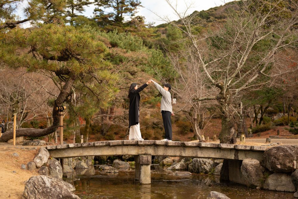 Couple dancing and holding hands on a stone arch bridge in a tranquil Japanese garden setting.