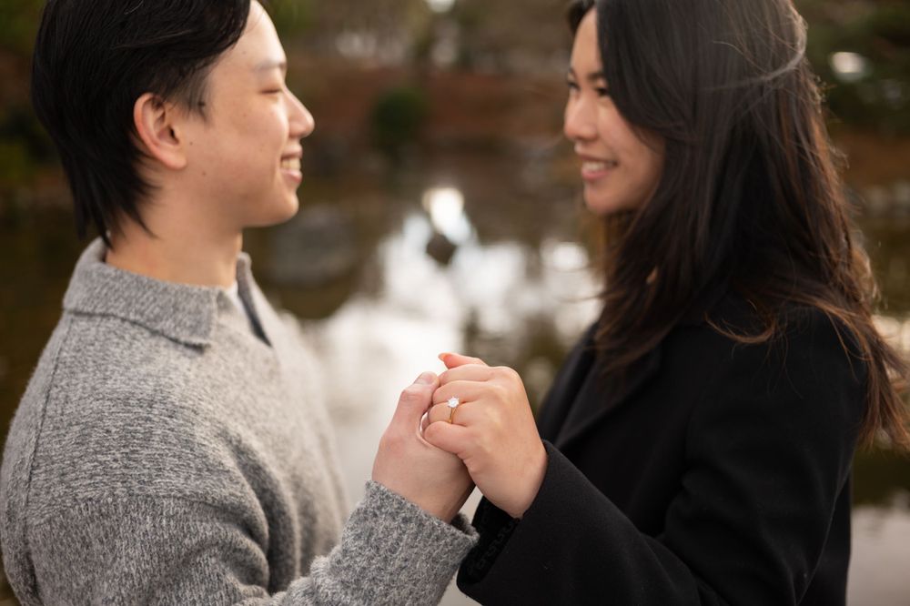 A couple holds hands and smiles at each other outdoors near a serene pond during an engagement session.