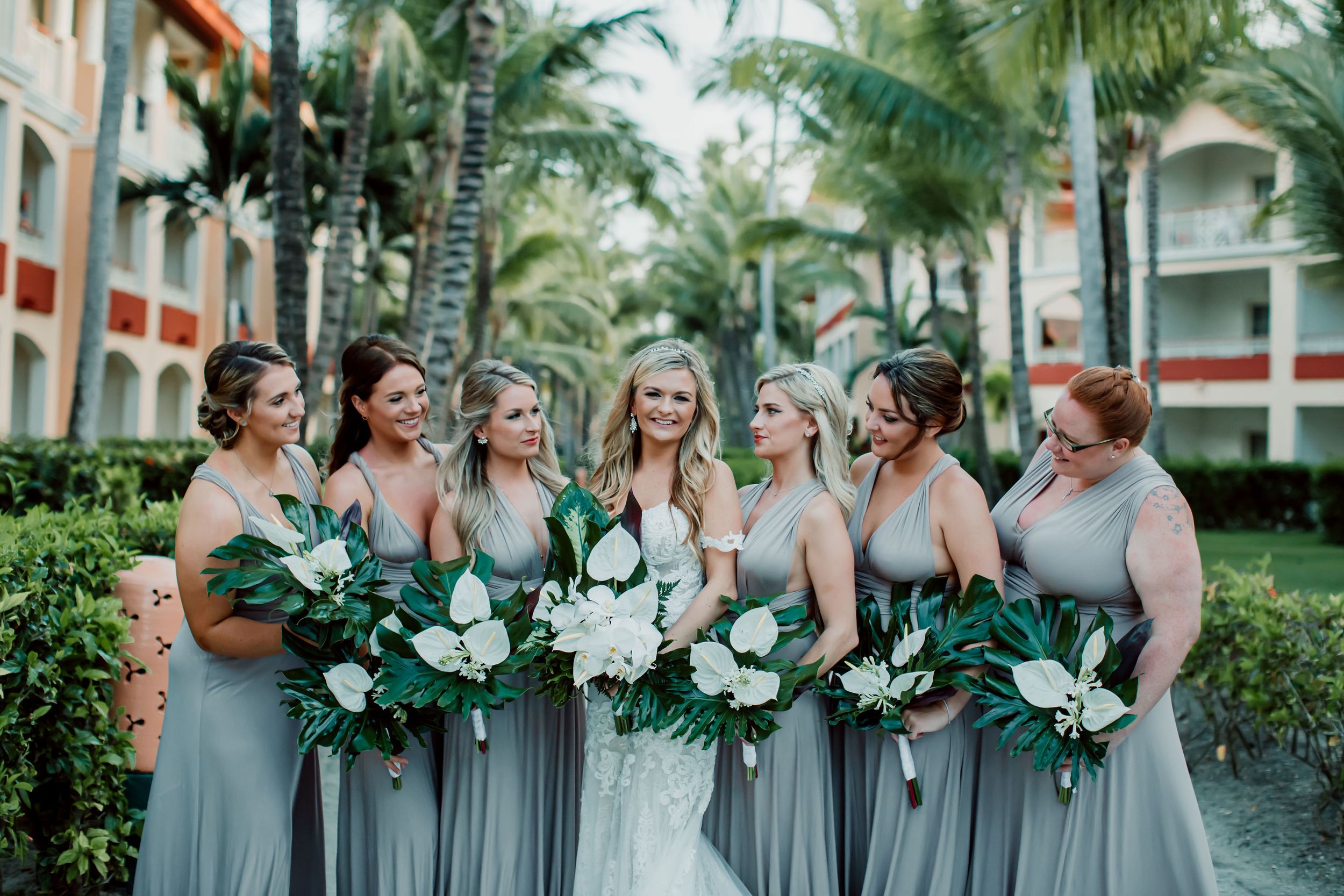 Wedding party in sage green dresses poses with floral bouquets among tropical palm trees.