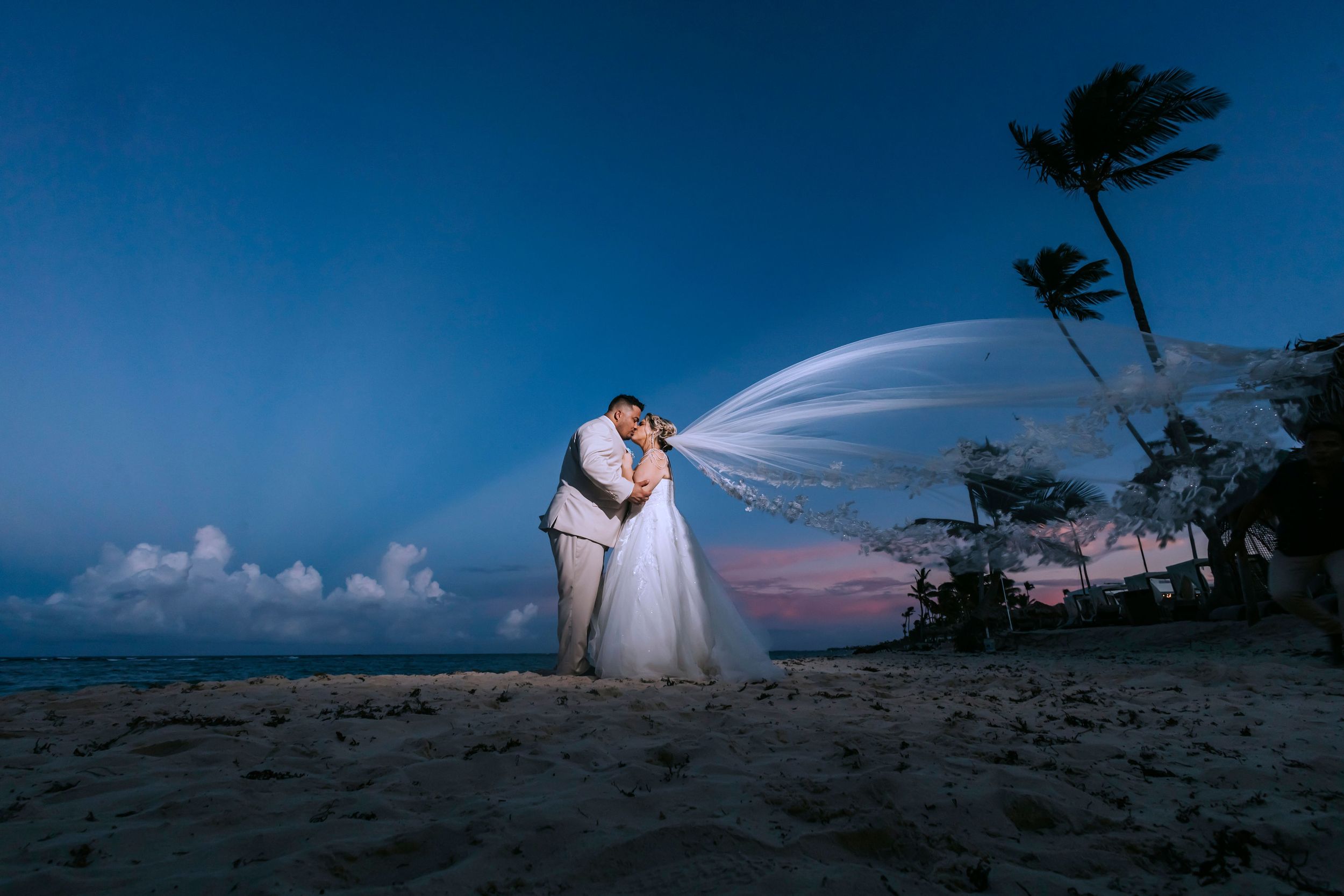 A romantic couple in wedding attire share a sunset moment on a windy beach with a flowing veil and palm trees against blue sky.