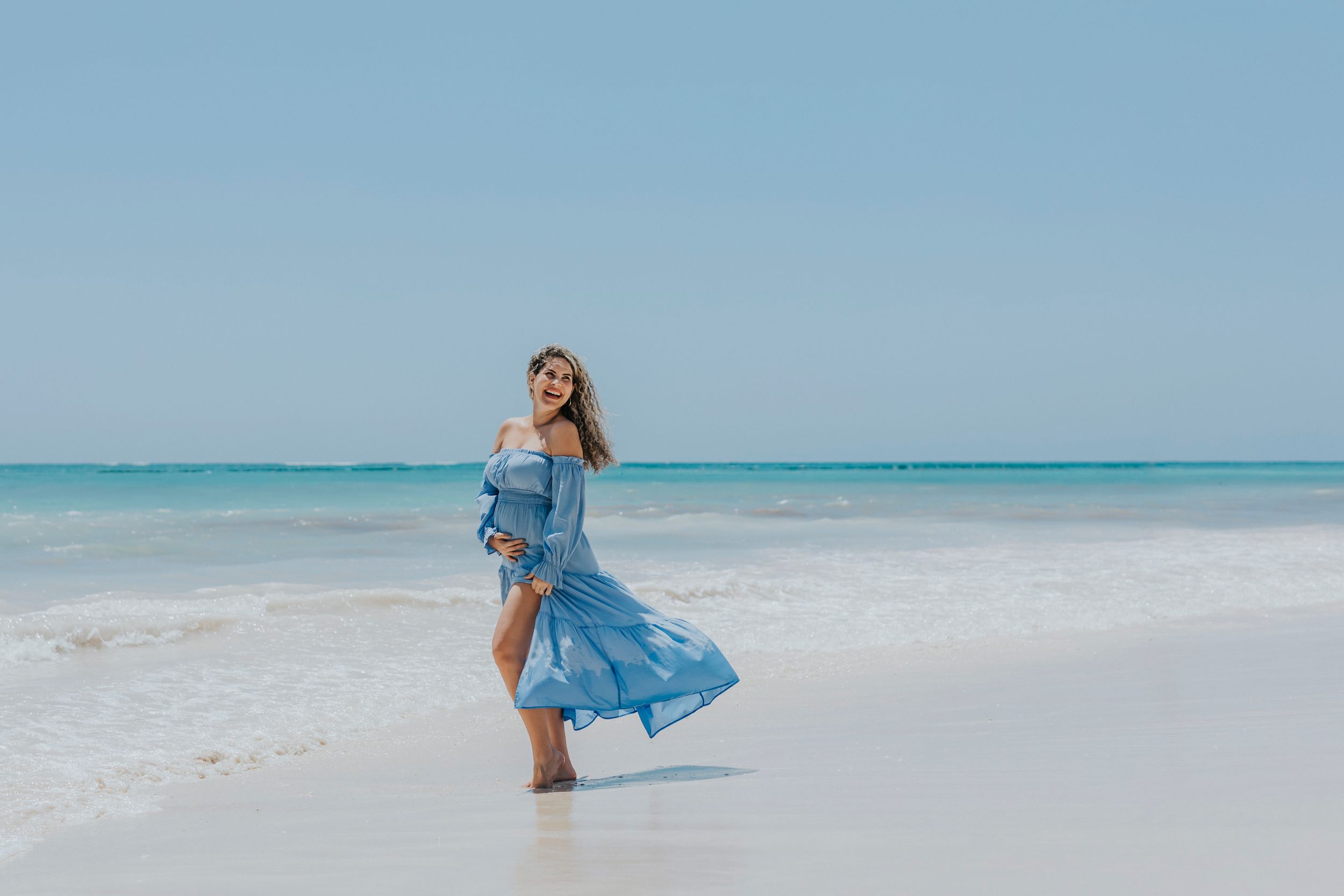A flowy blue dress catches the ocean breeze as someone dances on a pristine white sand beach with turquoise waters behind.