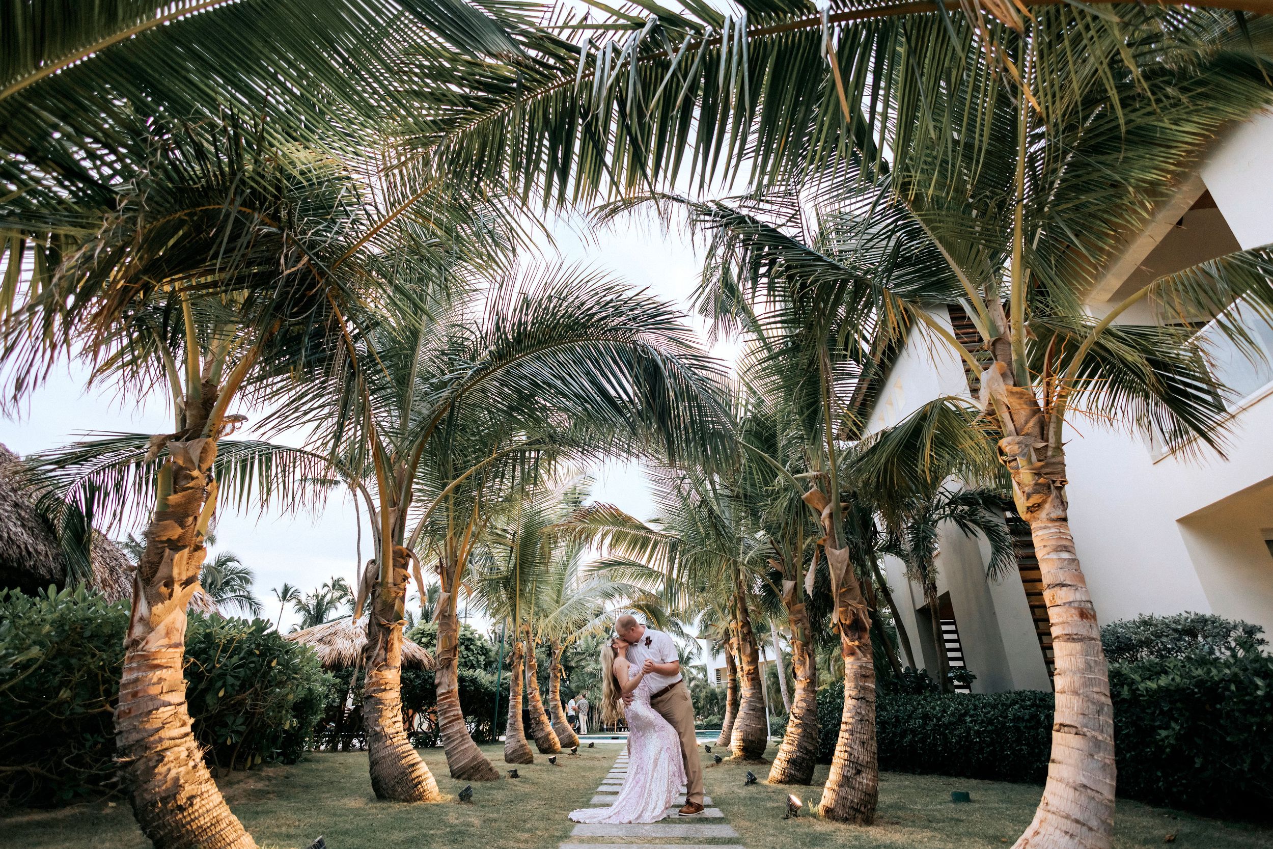 Elegant couple shares a romantic moment under palm trees lining a tropical pathway at sunset.