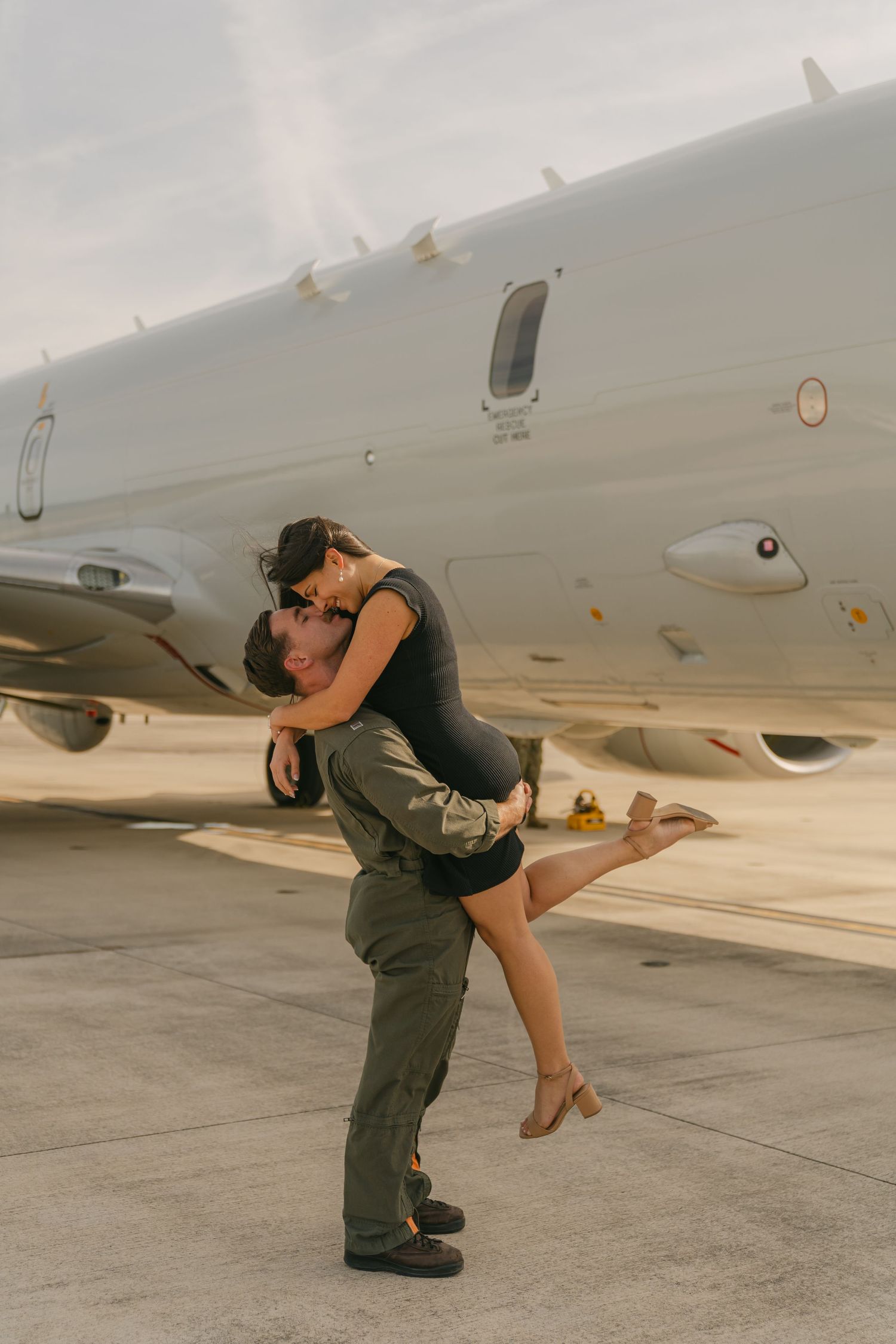 Military couple shares romantic moment next to aircraft on tarmac.
