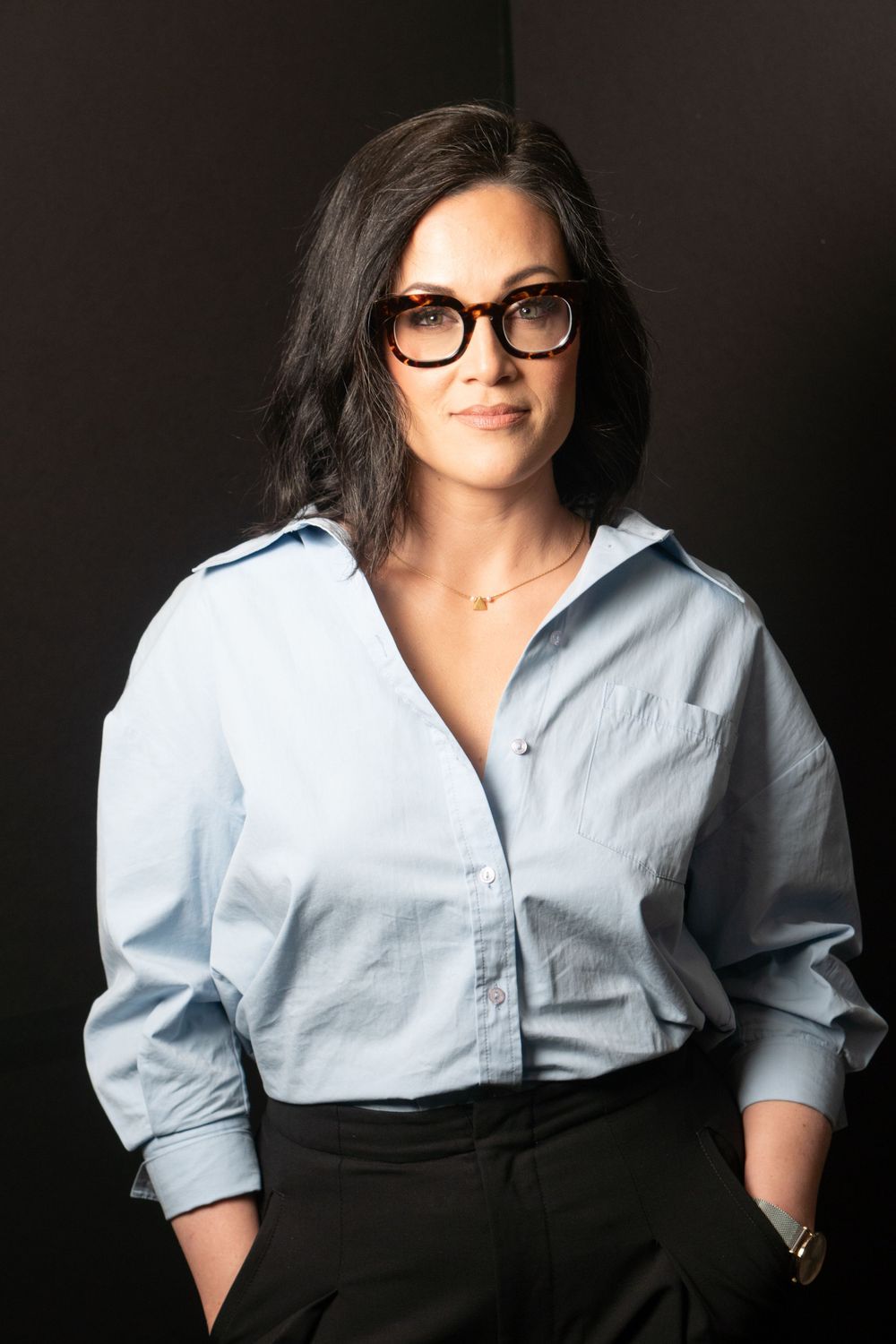 Business portrait in light blue button-down shirt and black pants against dark background, hands in pockets.