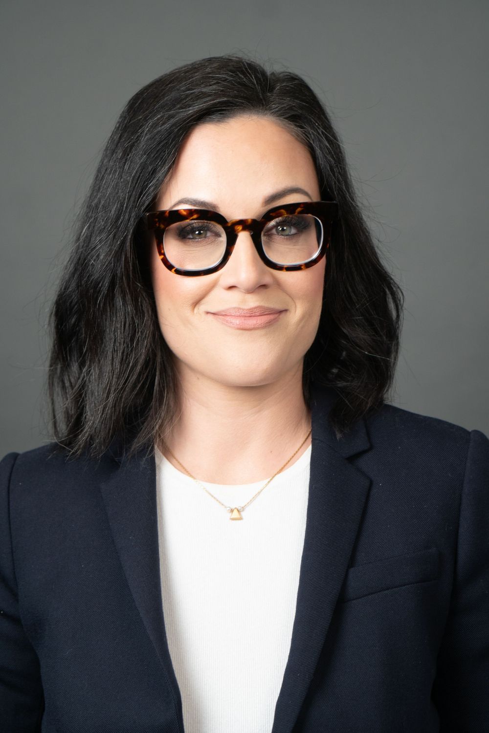 Close-up professional headshot with black blazer, white top and glasses against gray background.