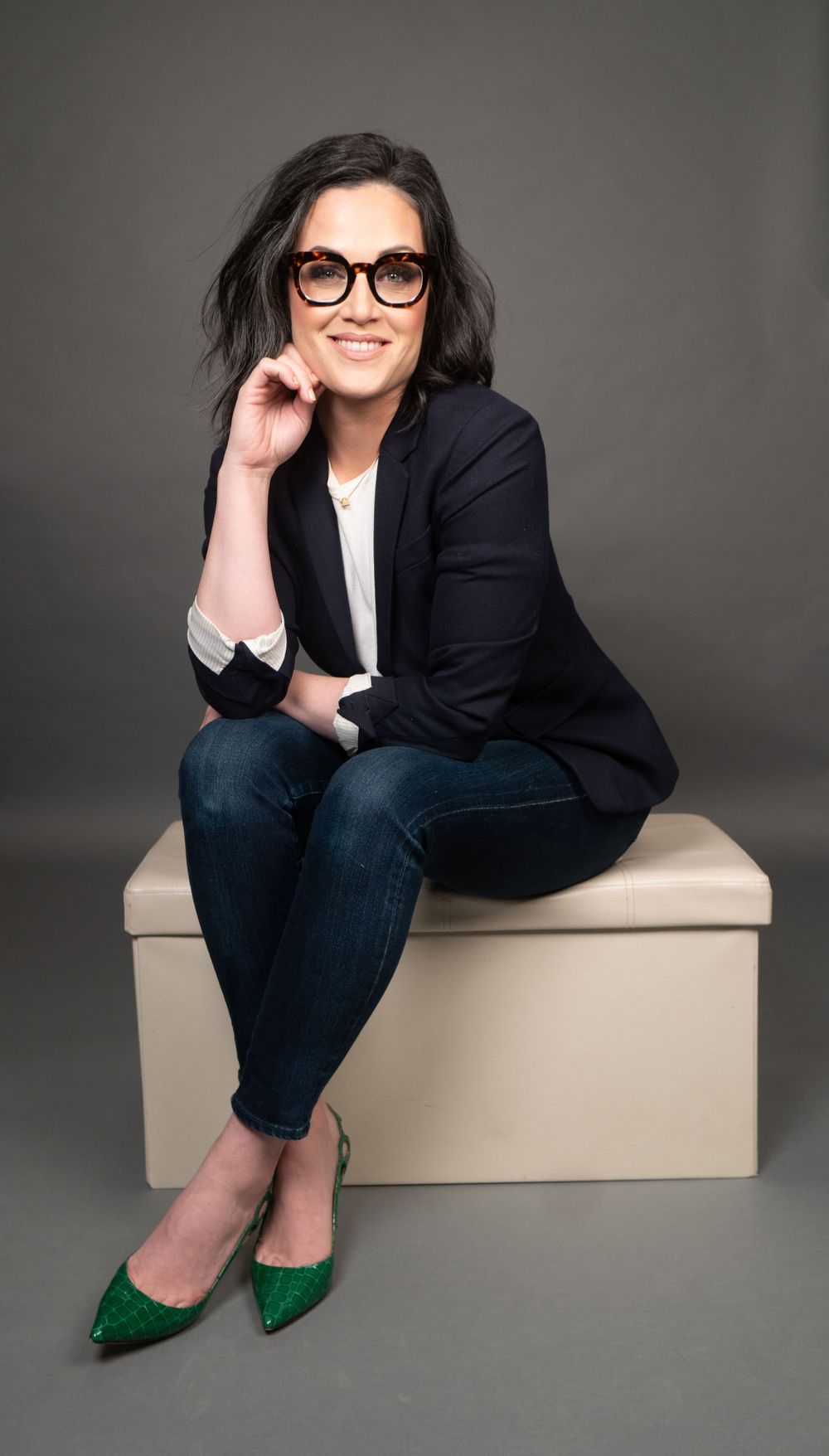 Professional portrait of person in black blazer and green shoes seated casually on beige cube against gray background.