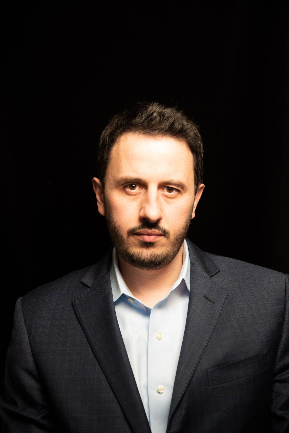 Close-up professional portrait of a person wearing a dark suit and light shirt against black background.