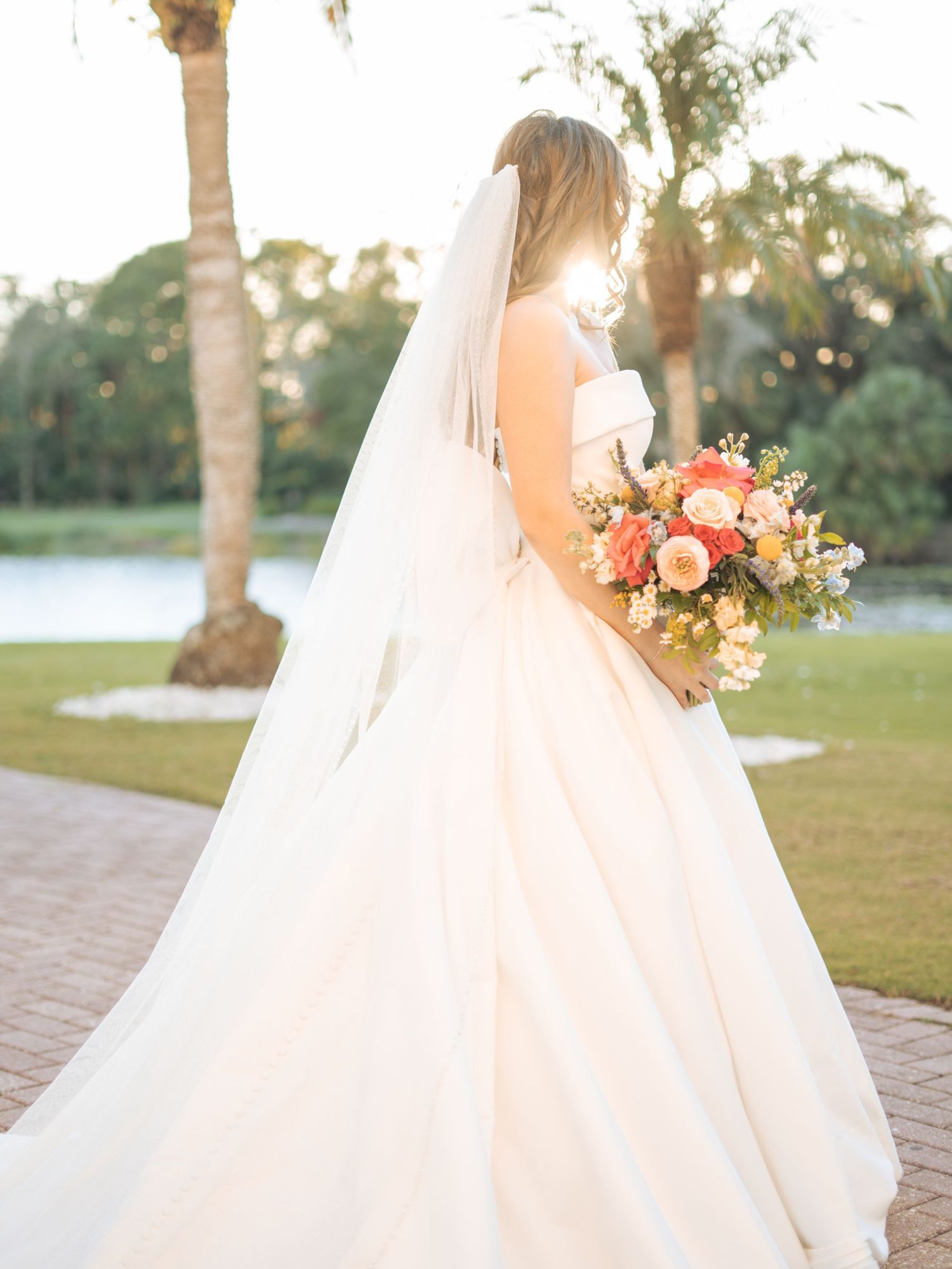 A bride in a flowing white gown holds a coral and peach bouquet while standing on a brick path near palm trees at sunset.