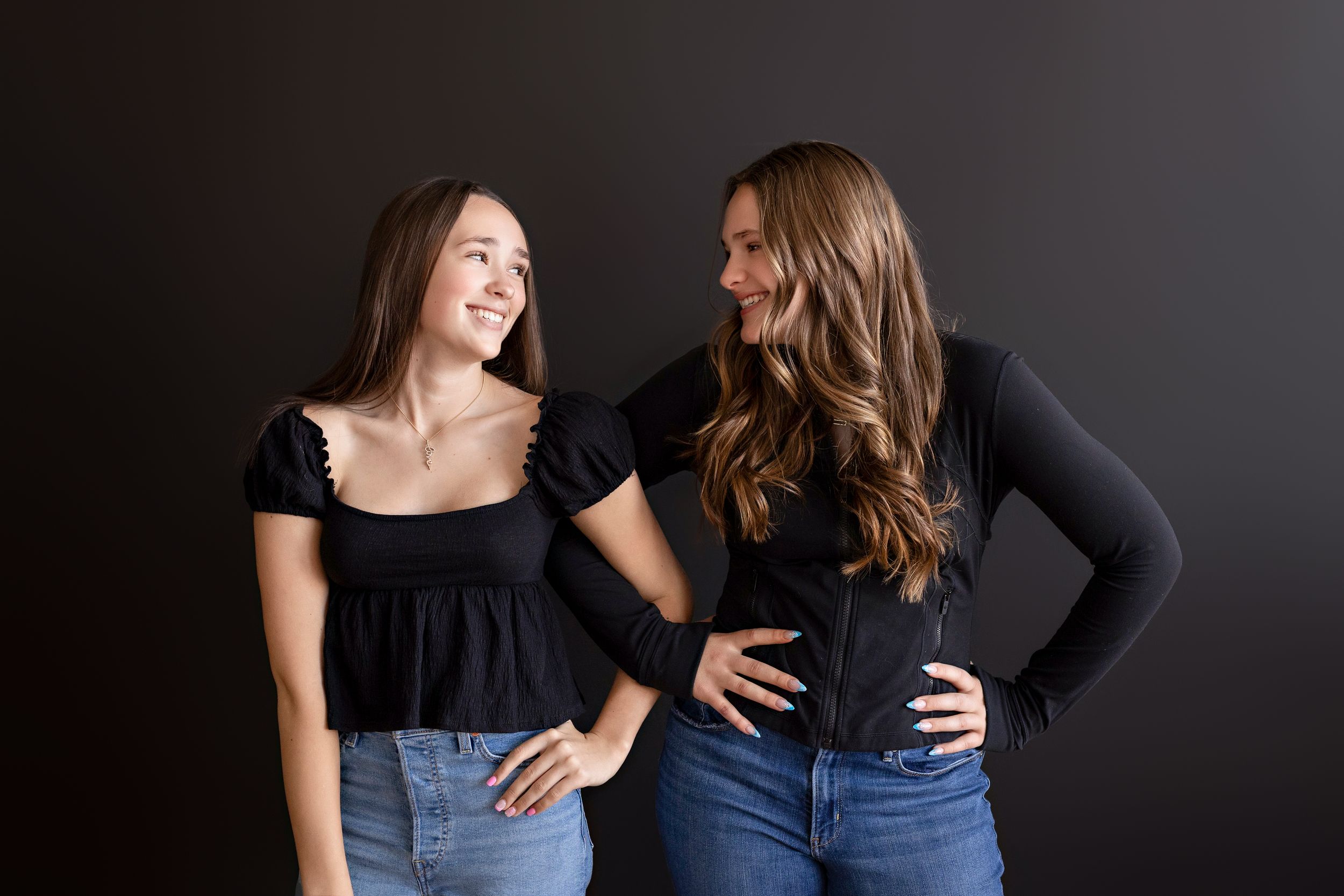 Two people in black tops and blue jeans pose with hands on hips against a dark background.