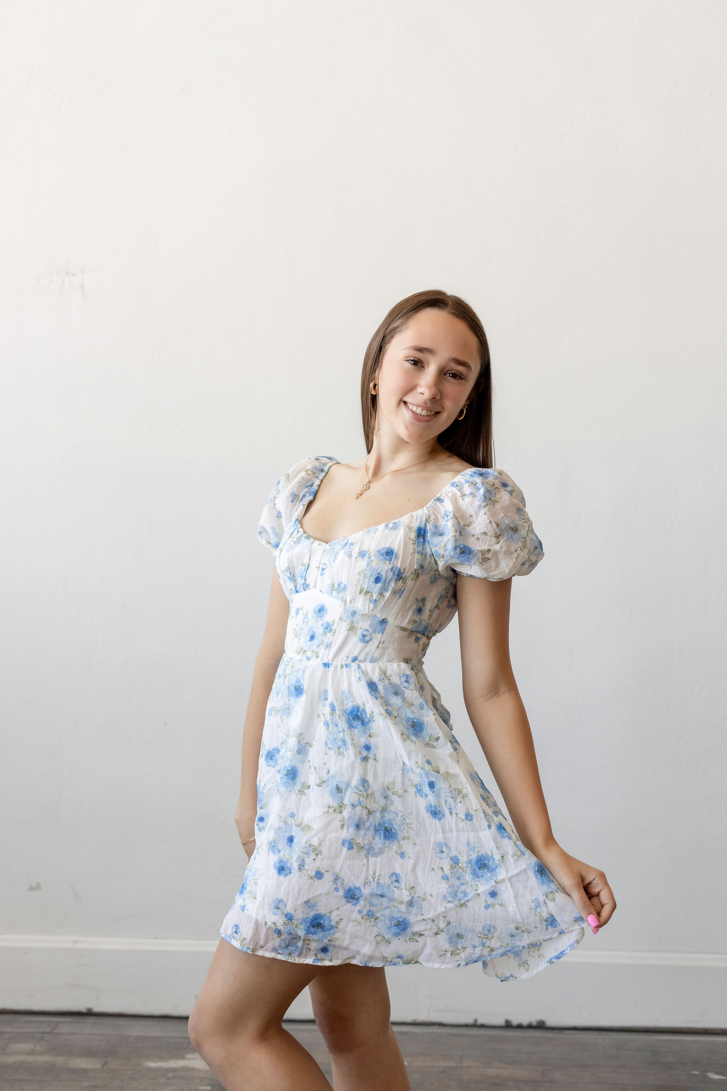 A young woman in a white and blue floral dress with puff sleeves poses against a plain white wall.