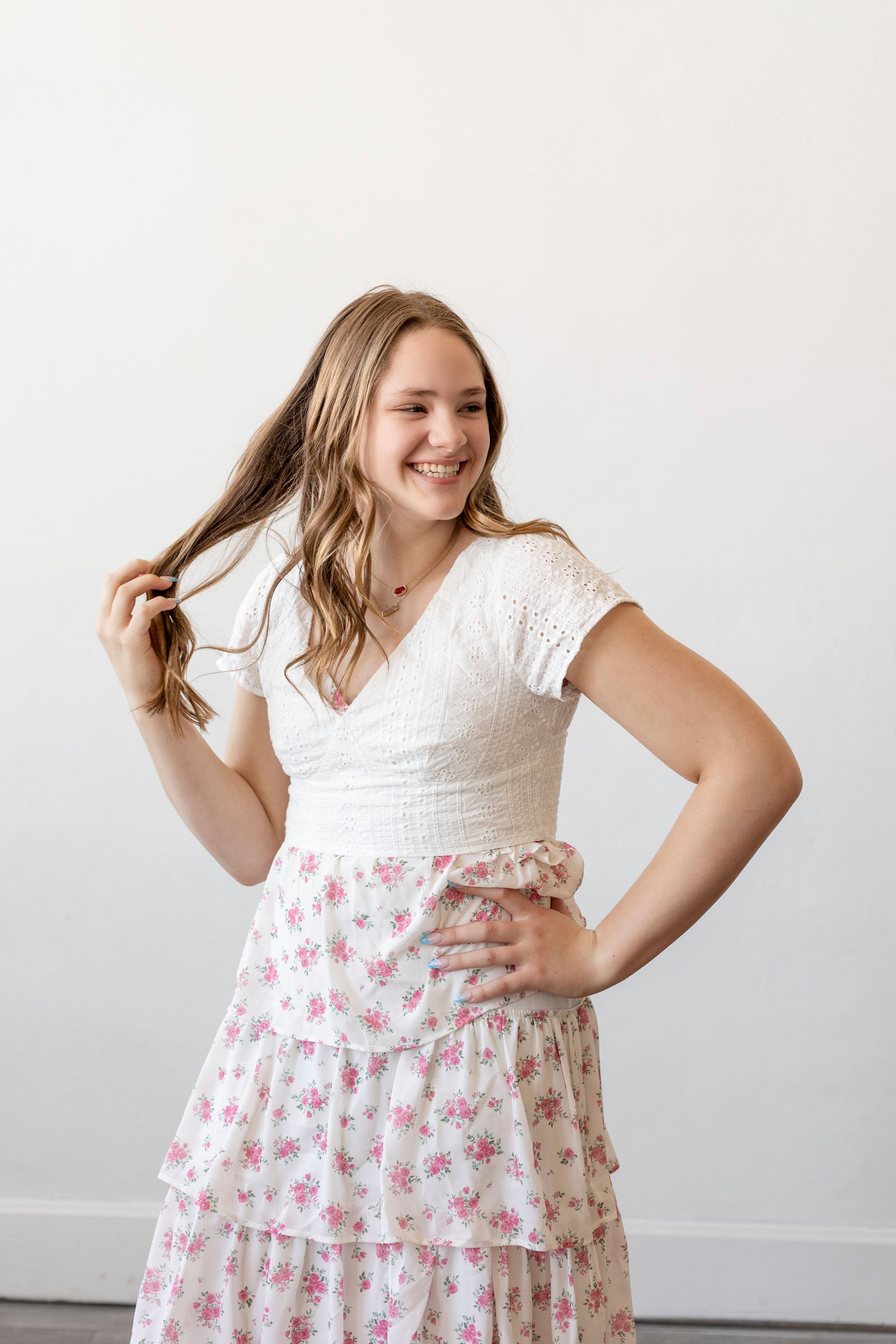 Young woman in white lace top and floral skirt stands against white wall while playing with her hair.