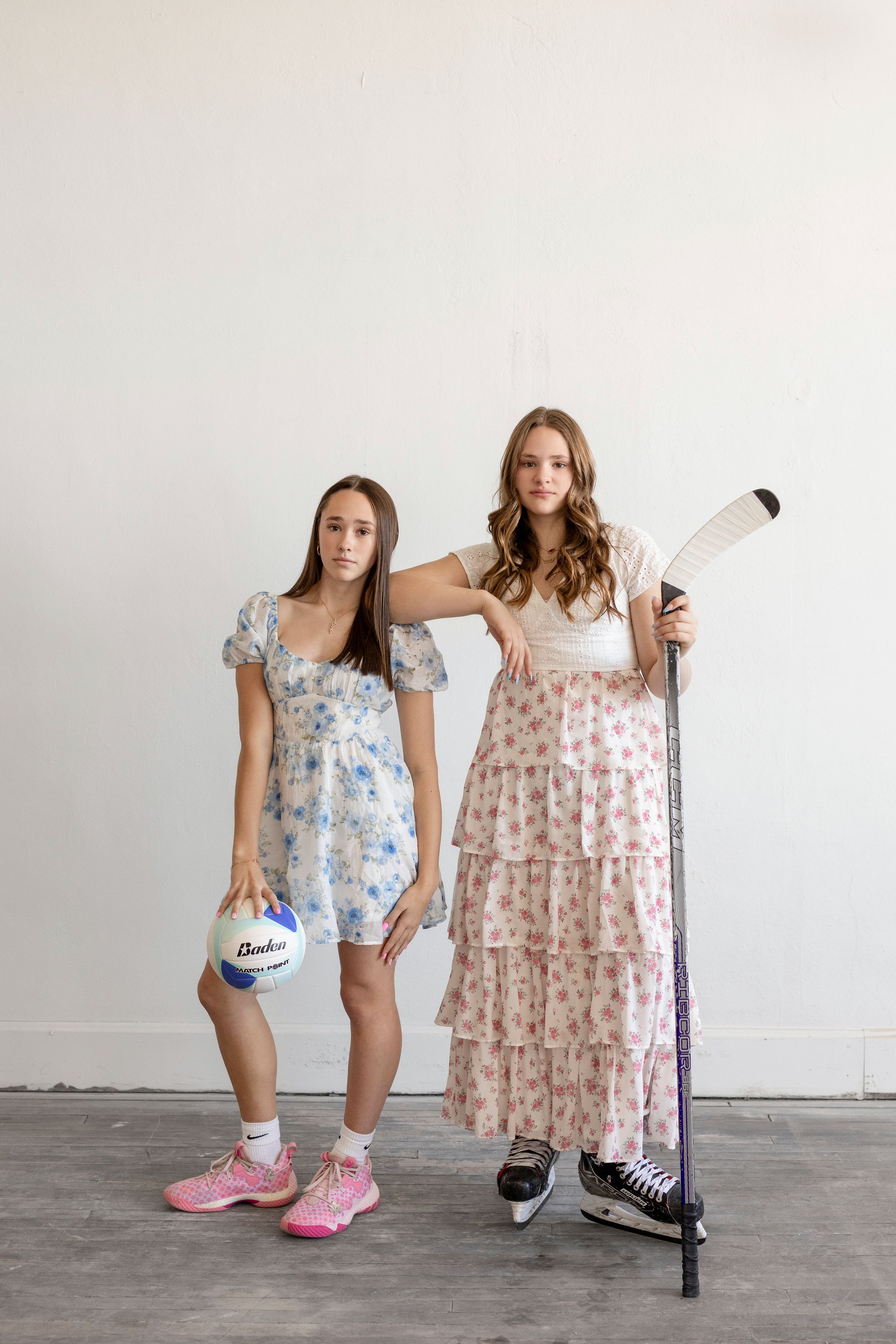 Two athletes in floral dresses pose with field hockey stick and volleyball against white wall.