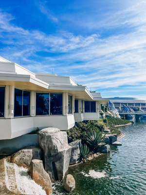 Modern waterfront building with white exteriors and large windows perched on rocky cliffs above blue waters on a sunny day.