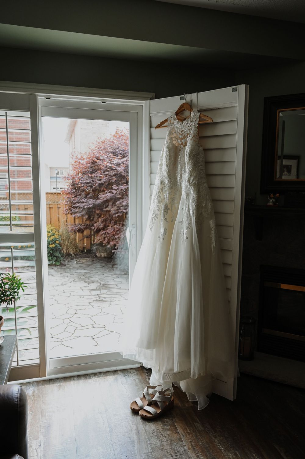 An elegant white lace wedding dress hangs by a bright window with wedding shoes beneath on hardwood floors.