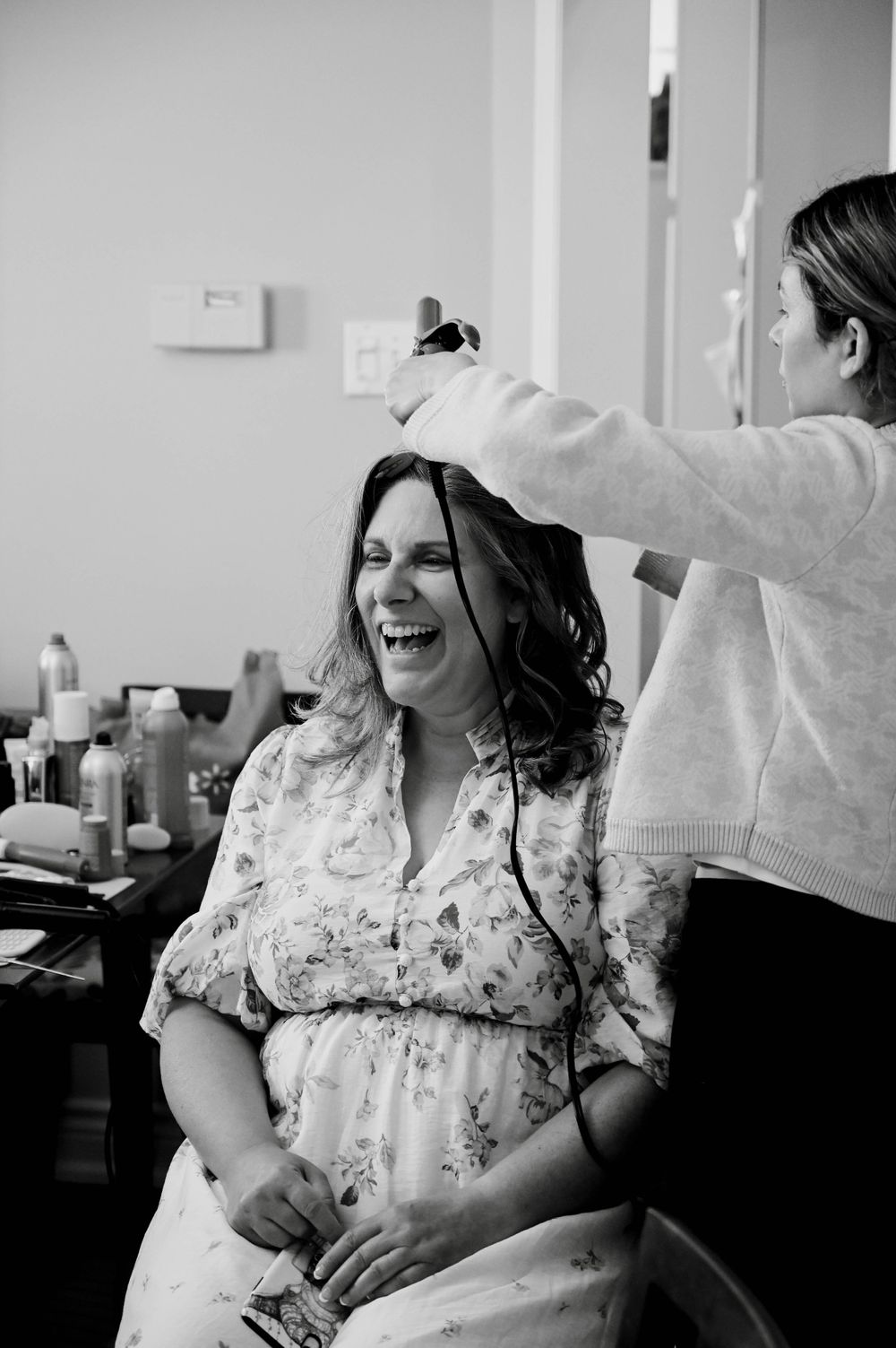 Black and white photo of someone getting their hair styled and laughing while wearing a floral dress in a salon setting.