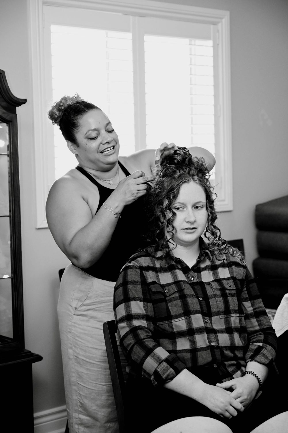 A hairstylist styles a client's curly hair while working in a bright, window-lit room.