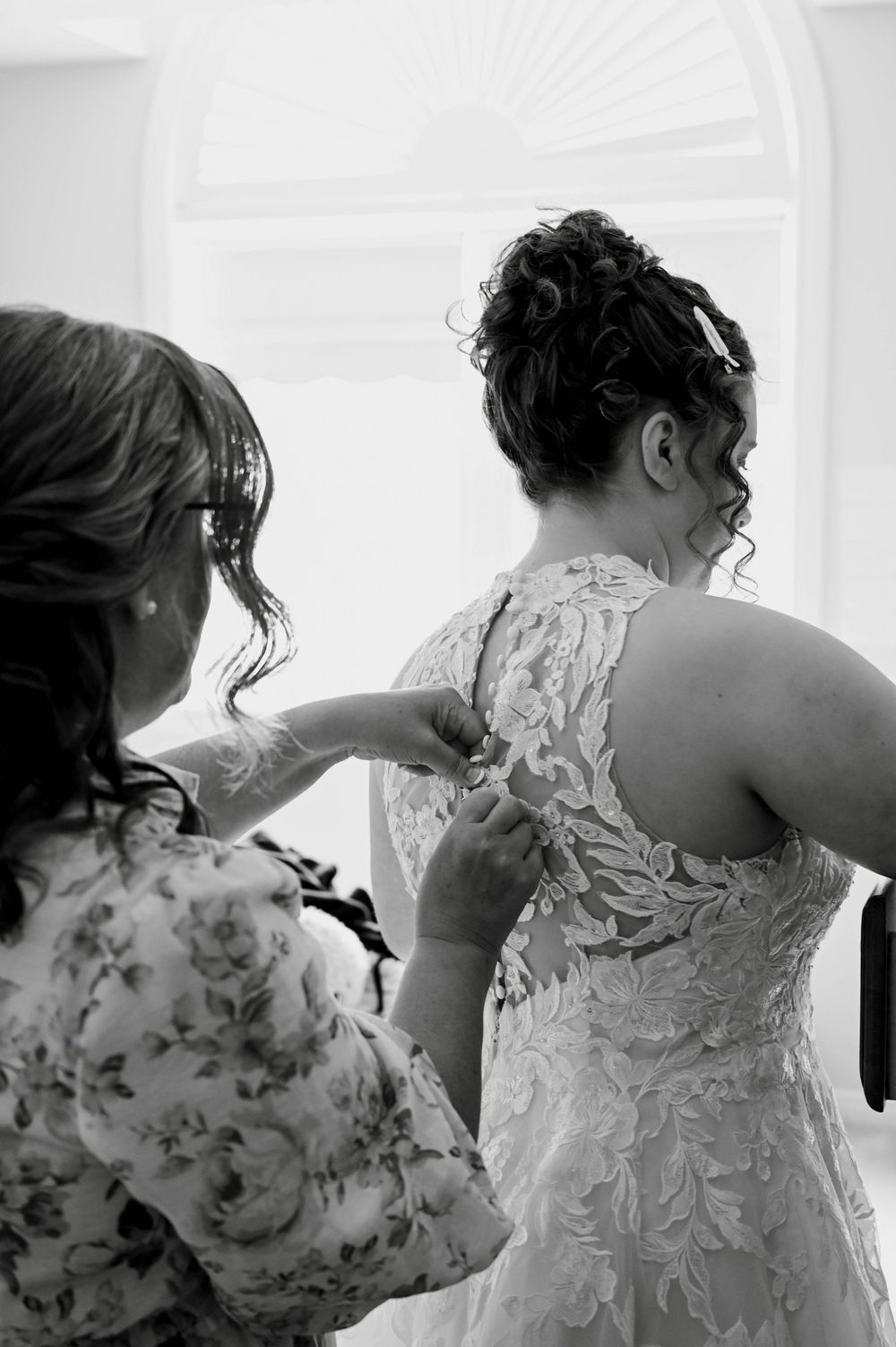 Black and white photo showing the intricate lace detail of a wedding dress being buttoned up during bridal preparations.