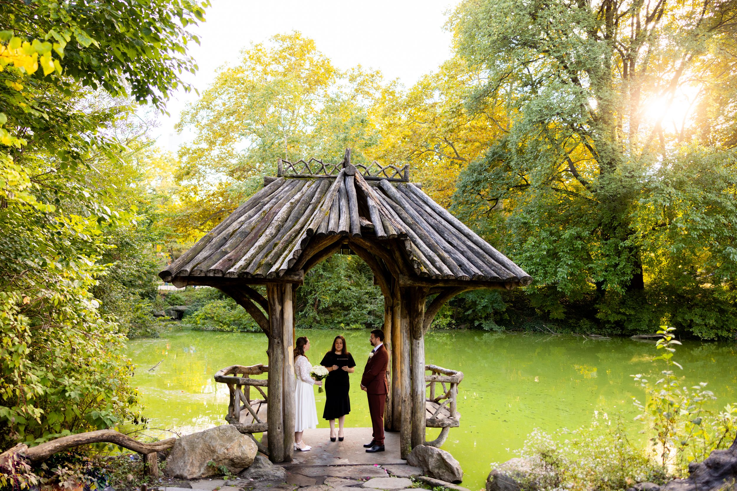 A silhouetted couple stands in Wagner Cove NYC Central Park a rustic wooden gazebo surrounded by green foliage.
