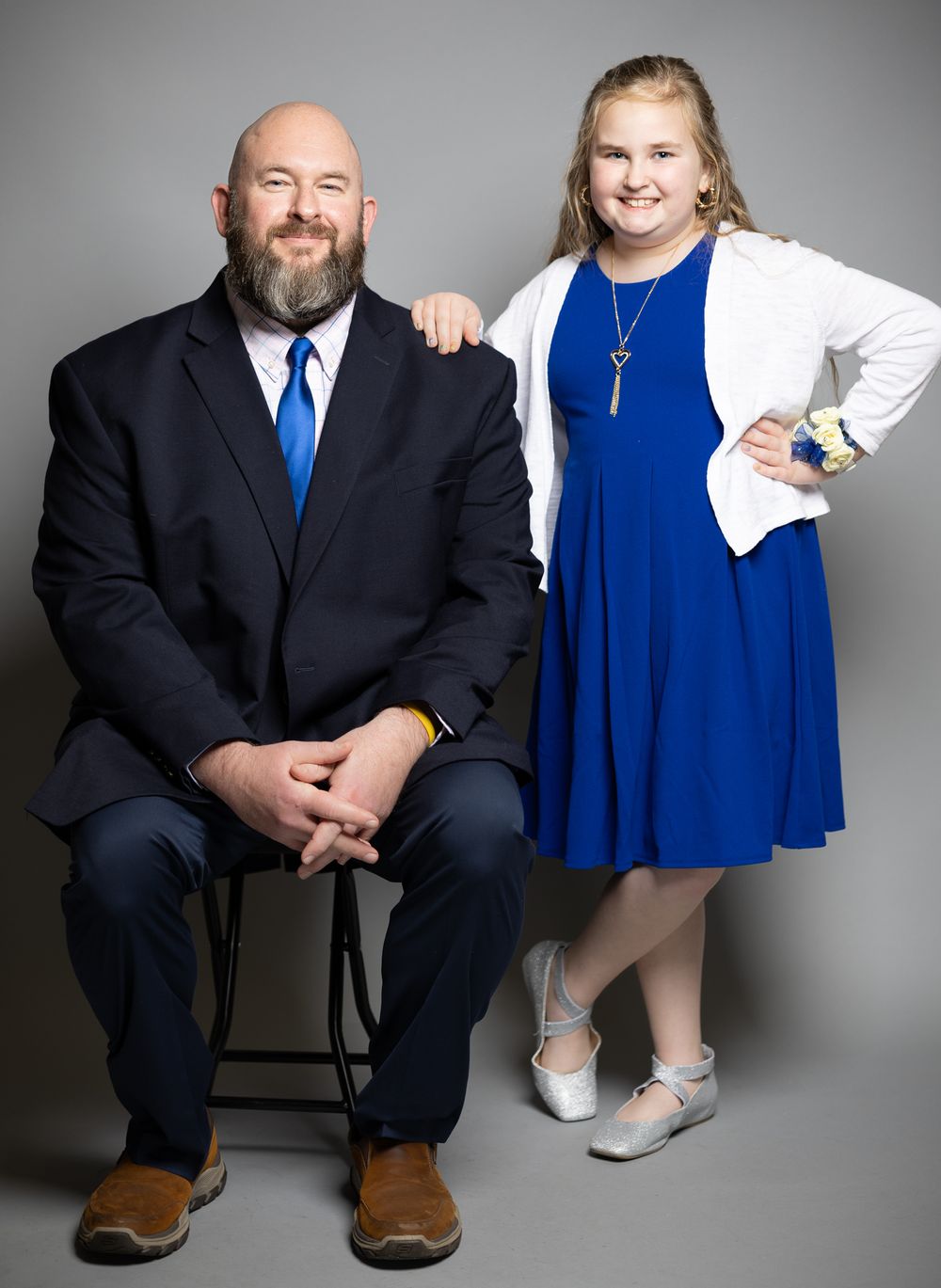 Studio portrait featuring adult in black suit and child in blue dress against grey backdrop.