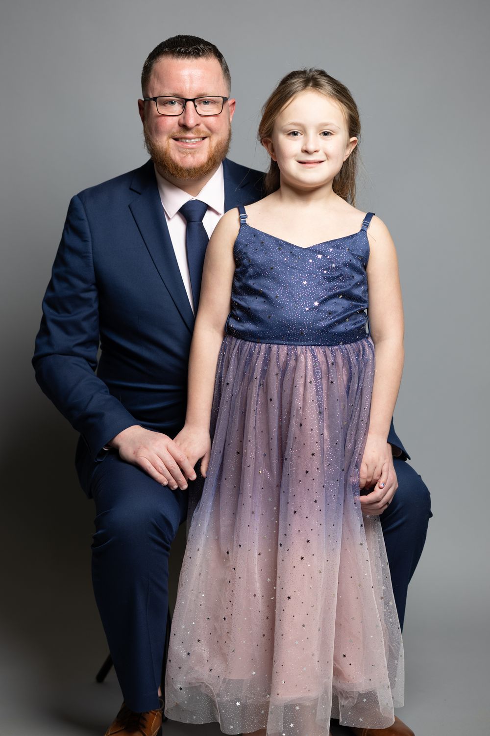 Formal portrait with adult in navy suit and child in sparkly purple dress against grey background.