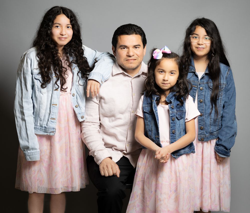 Family portrait with four people wearing coordinated pink and denim outfits against grey background.