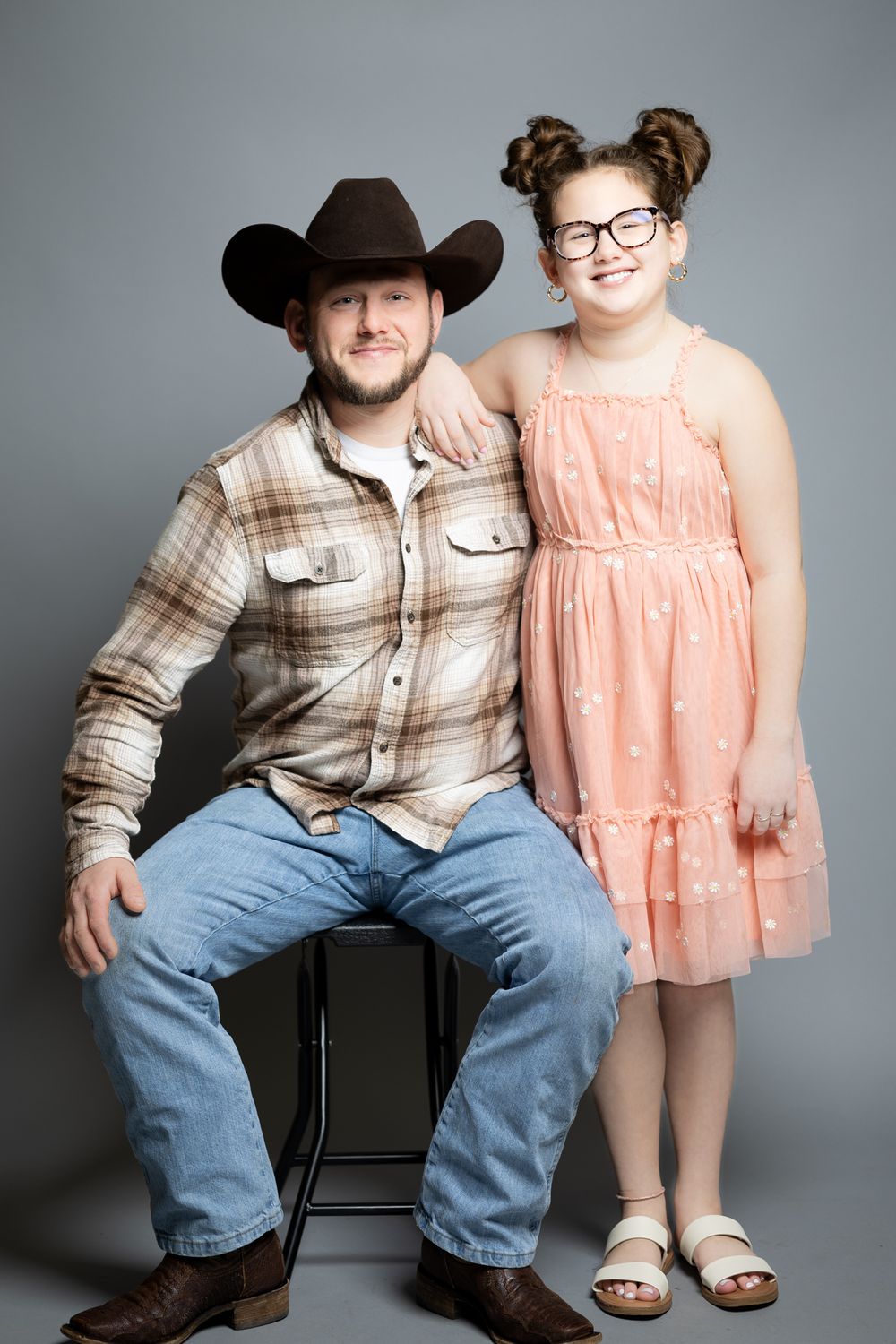 Studio portrait with person in cowboy hat and plaid shirt beside child in peach dress.
