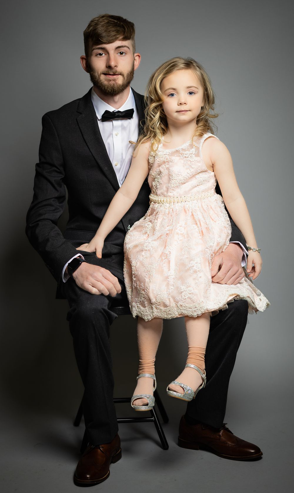 Studio portrait of two people in formal wear seated on a black stool against gray background.