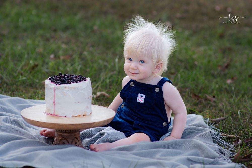 Toddler in navy overalls sits beside white cake with dark toppings on gray blanket outdoors.