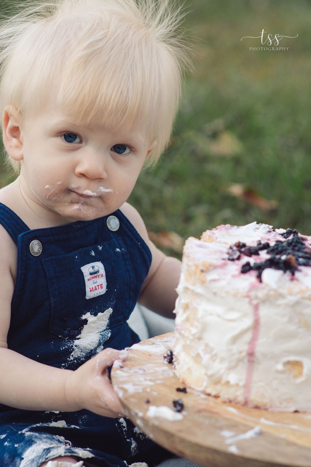 A toddler in blue overalls enjoys messy cake smash photography session outdoors.