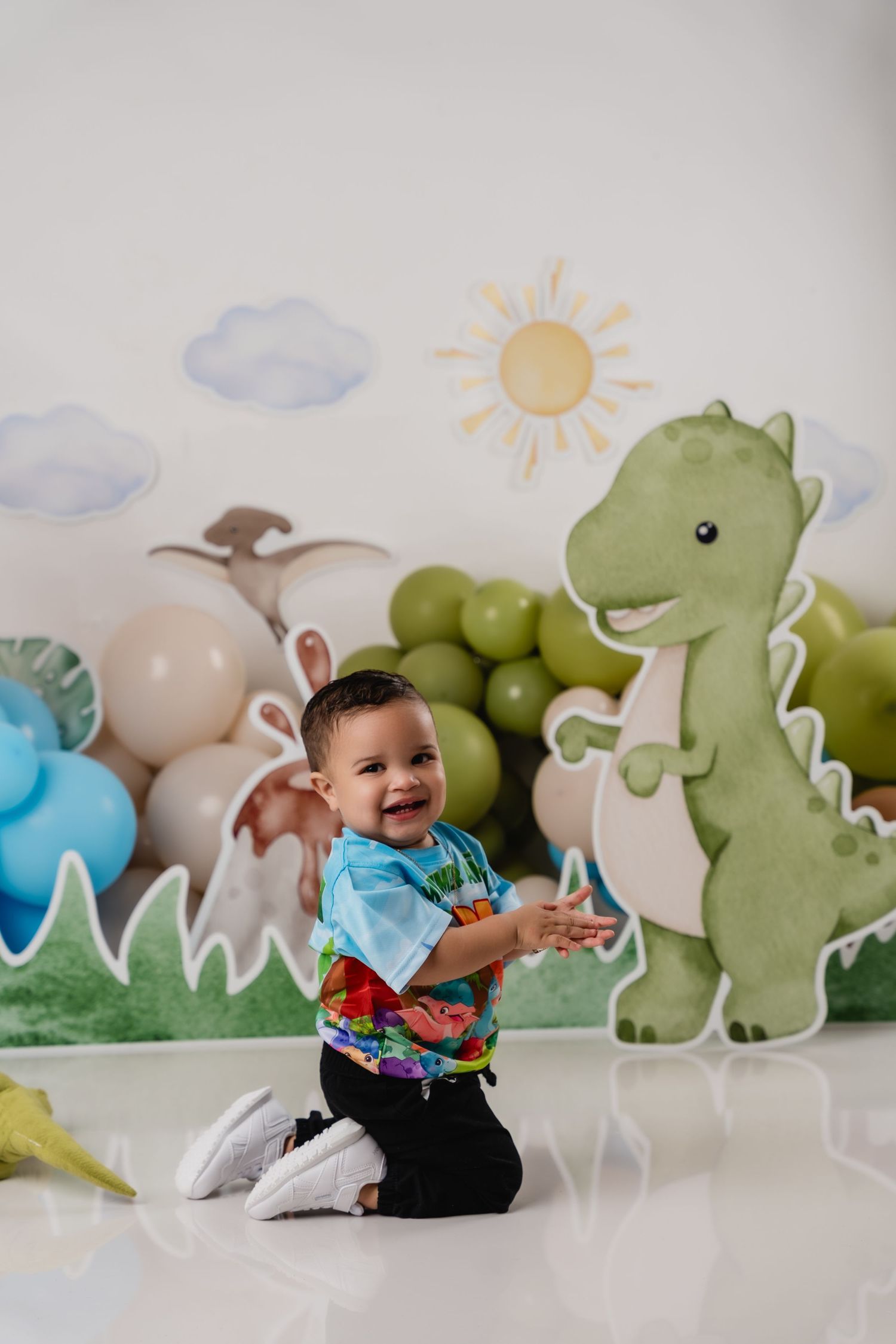 A dinosaur-themed party setup with balloons and a green cartoon T-Rex decoration against a painted wall with clouds and sun.