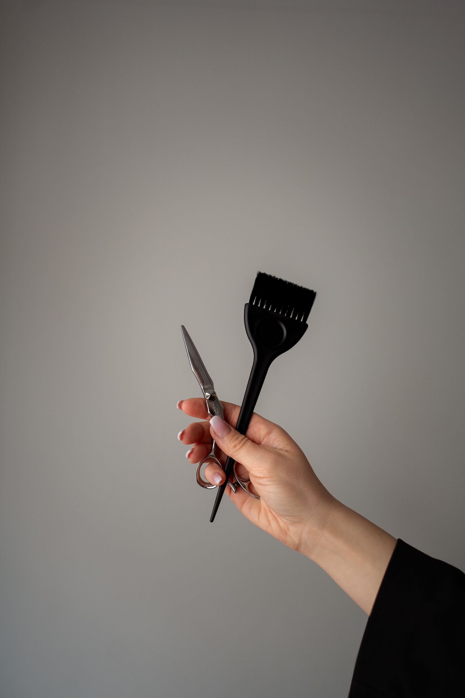 A hand holding hair cutting scissors and black tinting brush against a gray background.