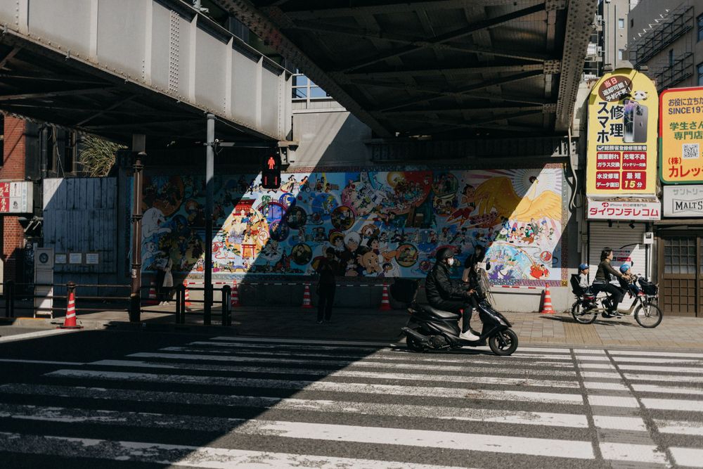 People crossing under a bridge with colorful graffiti in an urban Japanese setting.