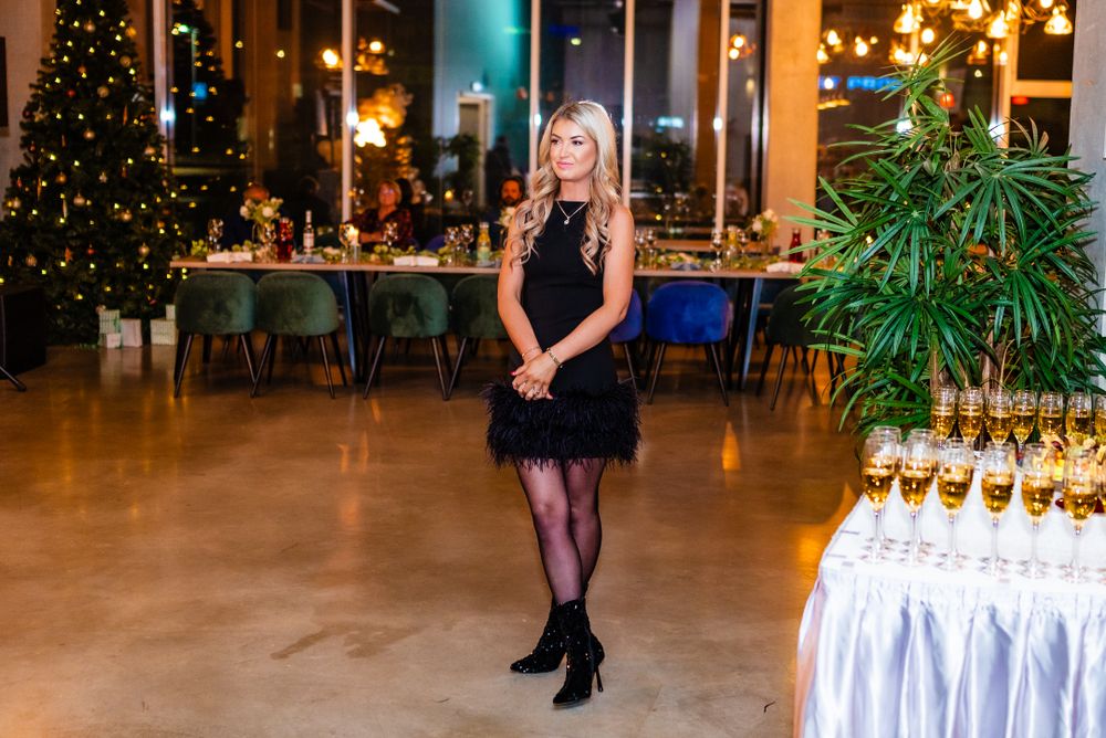 Woman in black cocktail dress poses near Christmas tree and champagne display at evening event.