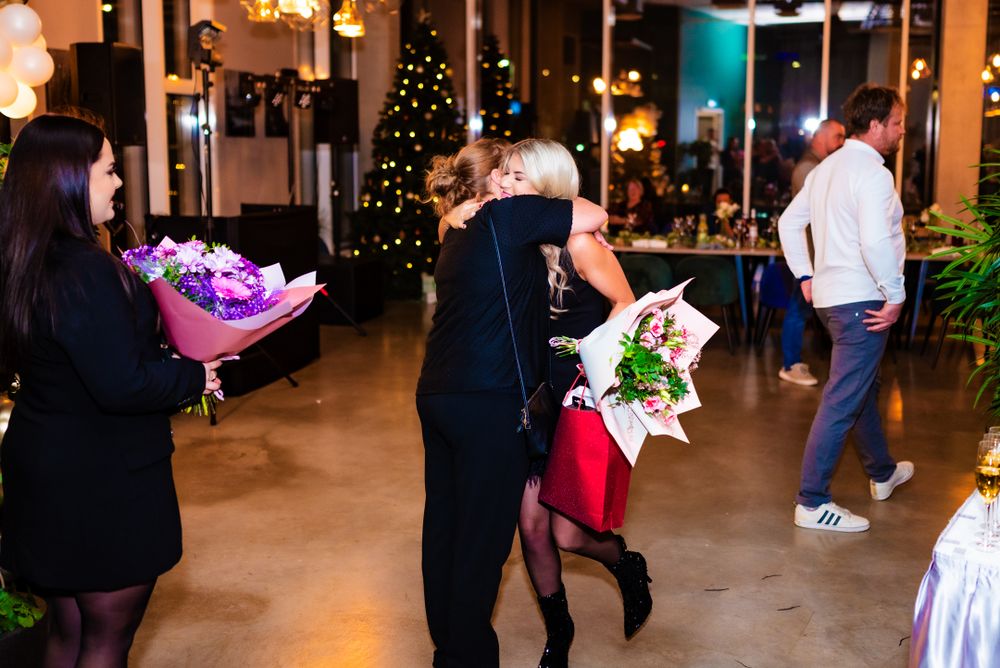 Party guests exchange flower bouquets at an evening celebration with holiday decorations.