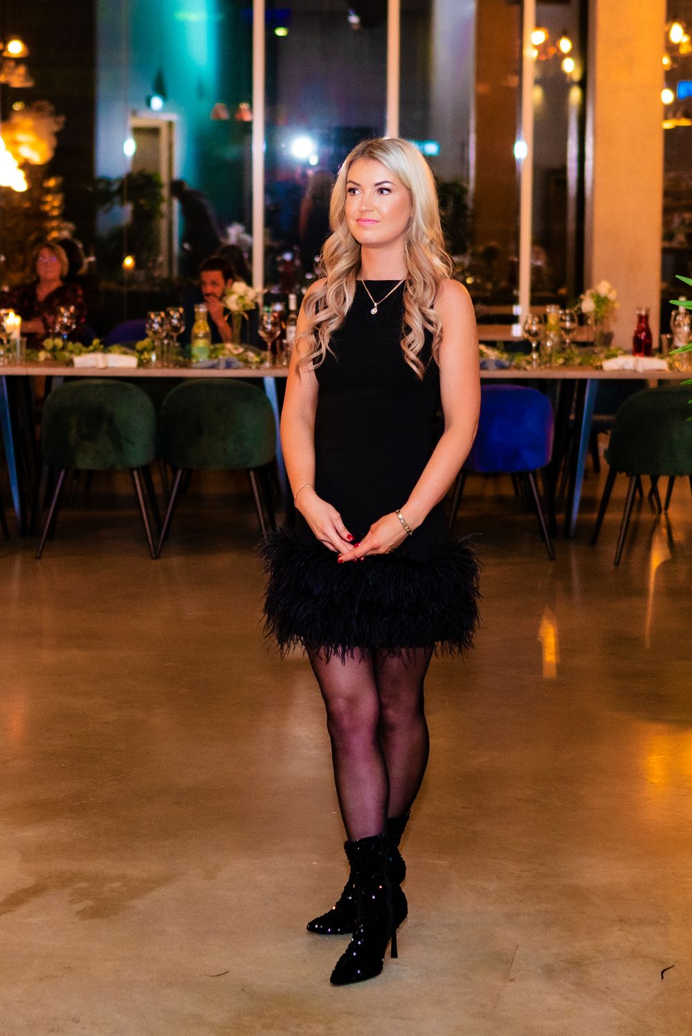 Woman in black feathered cocktail dress and tights poses at formal event with golden backdrop.