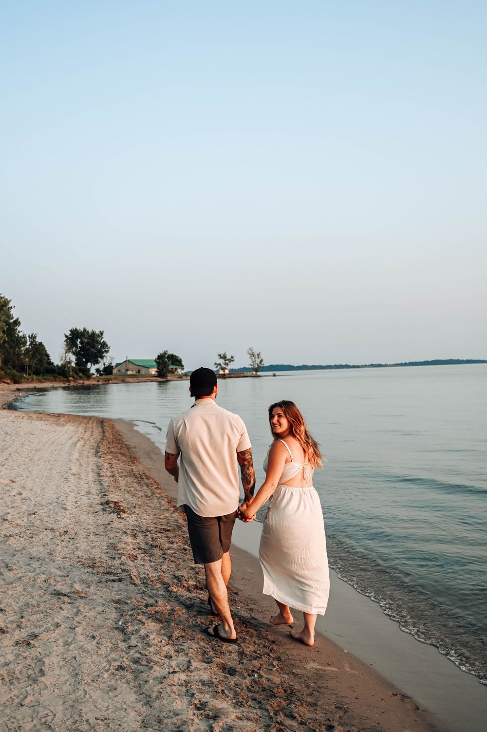 A couple walking hand in hand along a serene beach shoreline during sunset with calm waters and trees in the background.