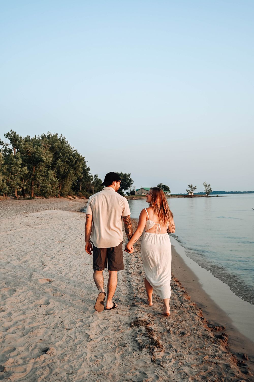 A couple walks hand in hand along a peaceful beach shoreline at sunset with trees in the background.