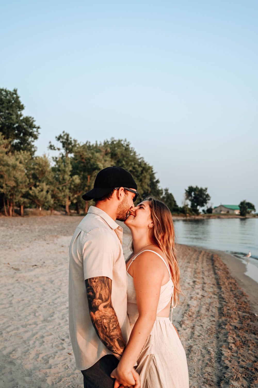 A romantic couple embracing and kissing on a sandy beach at sunset with trees and water in the background.