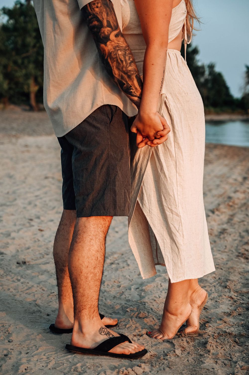Romantic couple holding hands on a beach at sunset with the person on left wearing shorts and right wearing a flowy dress.