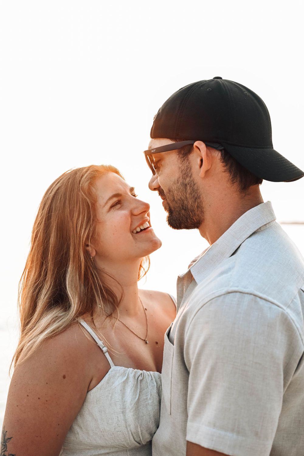 A romantic couple shares a tender moment laughing together at sunset, with one wearing a baseball cap.