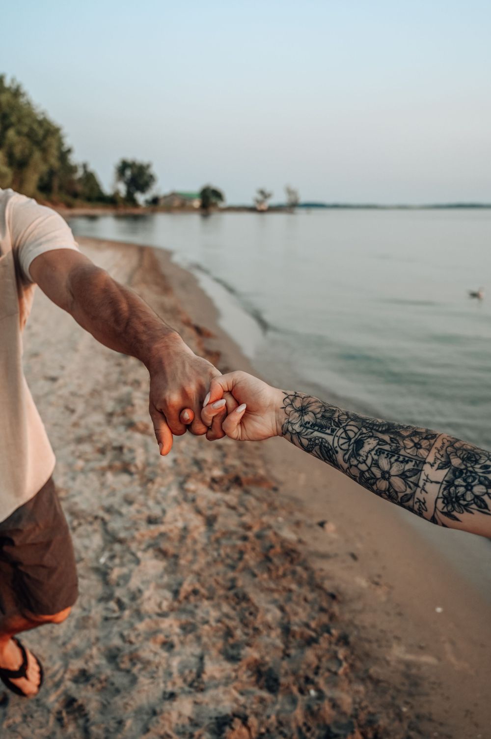 Two hands reach for each other on a beach shoreline with rocky sand and calm water in the background.